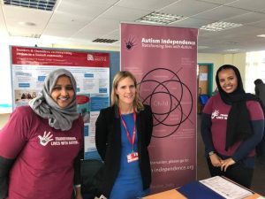 Three women stand in front of a notice board. Two of the women are wearing headscarves. All of them are smiling.