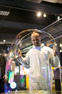 A man is holding a large bubble ring over their head. They have made a giant bubble that reaches the floor.